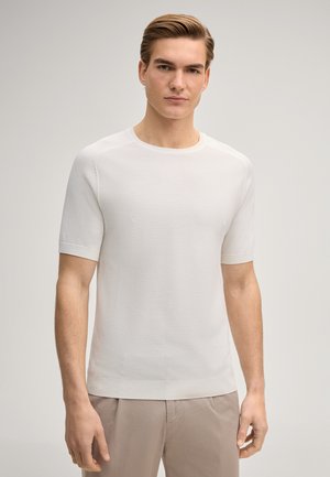 Junger Mann mit hellbraunem Haar, der ein weißes, strukturiertes Kurzarmhemd und hellbeige Hosen trägt und vor einem schlichten Hintergrund steht.