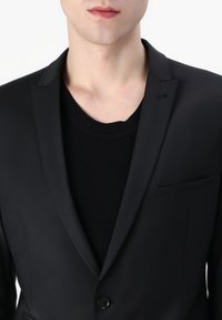 Zwarte blazer met puntige revers, enkelvoudige knoopsluiting en een borstzak, laag over een zwarte geribde trui. Glad stofoppervlak.