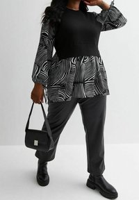 Frau trägt eine schwarze, gemusterte Bluse mit Puffärmeln, einen breiten schwarzen Gürtel, schwarze Hosen, klobige Stiefel und hält eine kleine schwarze Handtasche.