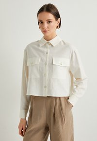 Camisa blanca de botones recortada en una tela ligera, con dos bolsillos en el pecho, cuello redondeado y botones tipo perla. Textura suave.