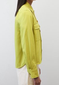 Jasnozielona kurtka zapinana na guziki z kołnierzykiem, kieszenią na piersi i mankietami. Materiał wydaje się gładki z delikatnym połyskiem.