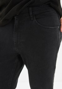 Närbild av en person som bär svarta denimjeans och en svart topp, med en hand vilande nära fickan.