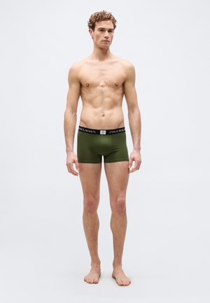 Mladić stoji bos, obučen u maslinasto zelene bokserice s crnim pojasom Polo Ralph Lauren, na jednostavnoj bijeloj pozadini.