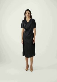 Μαύρο μίντι φόρεμα με σχέδιο περιτυλίγματος, κοντά μανίκια και λεπτομέρεια κόμπου στη μέση. Απαλή υφή, σε συνδυασμό με ανοιχτό μπεζ τακούνια.