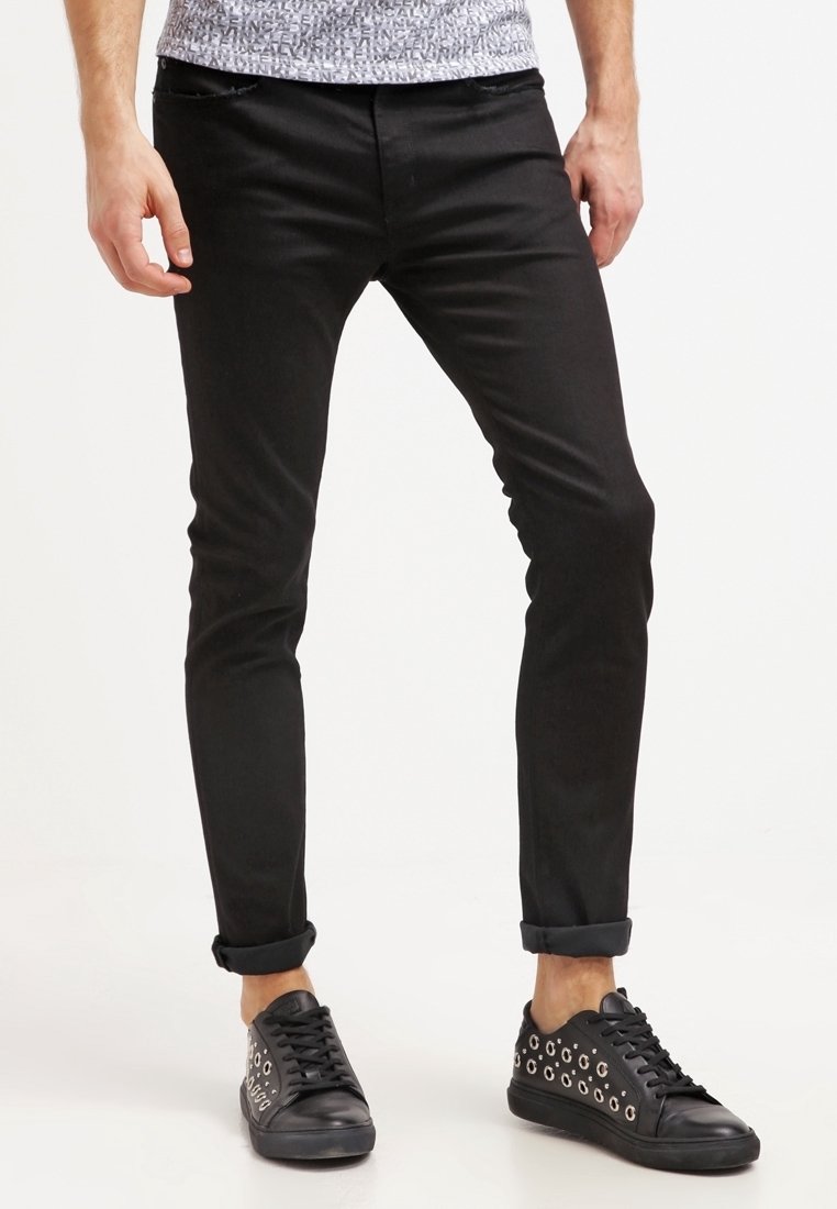 Homme portant un pantalon noir slim retroussé aux chevilles et des baskets noires avec des détails d'œillets argentés, se tenant devant un fond blanc.