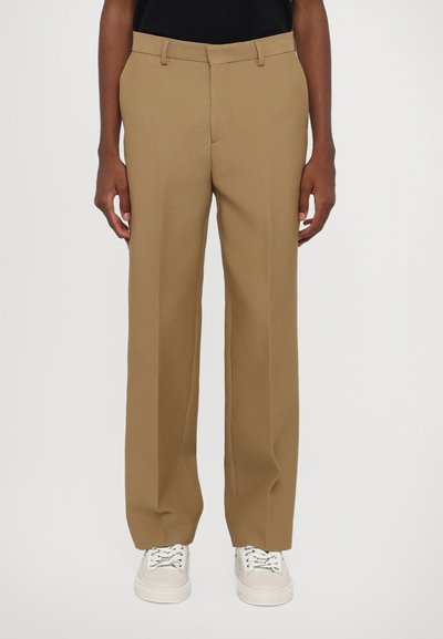 Μπεζ παντελόνι tailored με ίσια γραμμή, φτιαγμένο από λείο ύφασμα, με ζώνες για ζώνη και φερμουάρ, συνδυασμένο με λευκά αθλητικά παπούτσια.