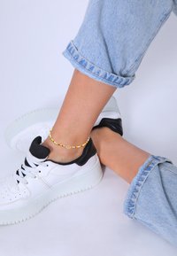 Perlekæde til anklen med farverige perler og et gyldent fredssymbol, båret sammen med opkrøllede lyseblå jeans og sorte og hvide sneakers.