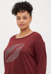 Top bordeaux de mangas longas feito de um tecido suave, com uma grande impressão metálica de lábios na frente. Apresenta um decote amplo e um corte descontraído.