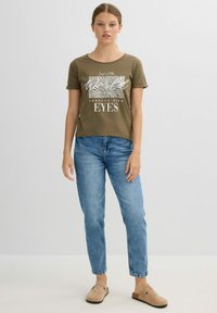 Camiseta verde oliva de manga corta y cuello redondo, con texto gráfico en blanco y líneas abstractas. Llevada con jeans azul claro y zapatos beige.