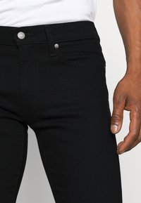 Jeans de mezclilla negra con un corte ajustado, que presentan un diseño clásico de cinco bolsillos y cierre de botón de metal. Textura suave y detalles de costura mínimos.