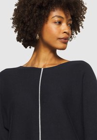 Marineblauer Strickpullover mit weitem Ausschnitt, der einen vertikalen hellgrauen Streifen auf der Vorderseite aufweist. Glatte Textur und lockere Passform.