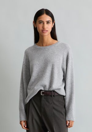 Vrouw met lang zwart haar die een lichtgrijze trui draagt, ingestopt in een donkergrijze broek met een donkerbruine riem, staand tegen een neutrale achtergrond.