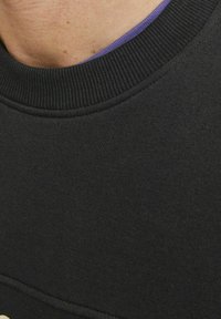 Sudadera negra con un cuello redondo acanalado y una textura suave. Una capa morada es visible debajo del cuello.