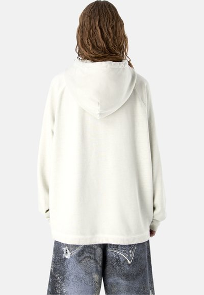 Personne aux longs cheveux portant un sweat à capuche blanc uni et un jean ample et usé de couleur foncée, vue de dos sur un fond uni.