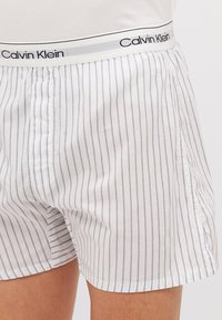 Calvin Klein muški bokserice u bijelom pamuku s tankim plavim vertikalnim prugama. Sadrži elastični pojas s logotipom i bočne proreze za udobnost.