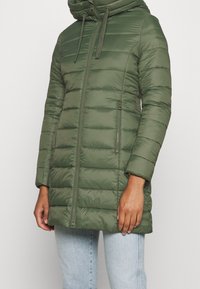 Abrigo acolchado de color verde oliva con cierre de cremallera en el frente, capucha y bolsillos laterales. Presenta costuras horizontales cosidas y una textura ligera acolchada.