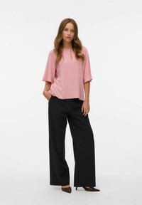 Donna in piedi con una blusa rosa a maniche corte con dettaglio a chiave, pantaloni neri a gamba larga e tacchi a punta marroni con ritagli.