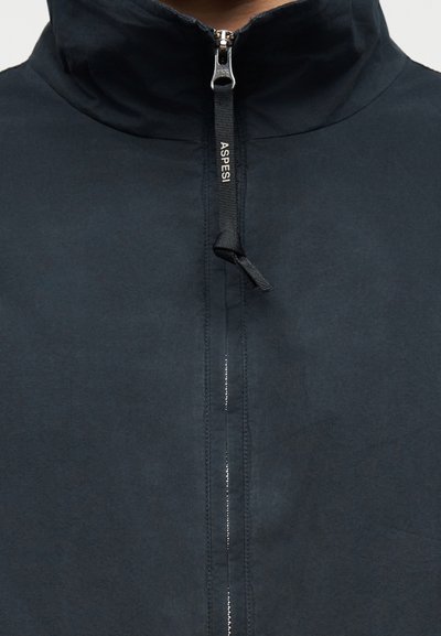 Κοντινό πλάνο γιακά μαύρου μπουφάν με ασημένιο φερμουάρ που διαθέτει μαύρη ταινία έλξης με την ένδειξη "ASPESI" σε λευκό κείμενο.