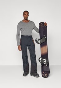 Gray long-sleeve top, black snow pants with orange zipper accents, and black snow boots. Holding a dark snowboard with white bindings.