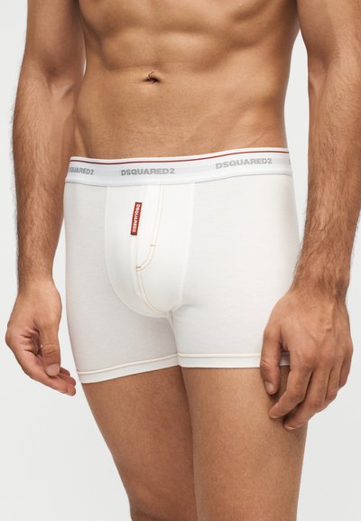 Torso masculin portant un boxer blanc DSQUARED2 avec une ceinture marquée et une étiquette logo rouge sur le devant sur un fond uni.
