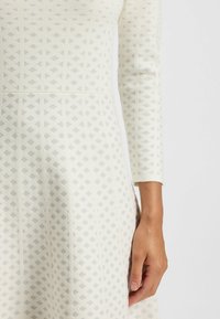 White dress featuring a textured diamond pattern in silver. Long sleeves with a fitted bodice and flared skirt. Smooth knit fabric.