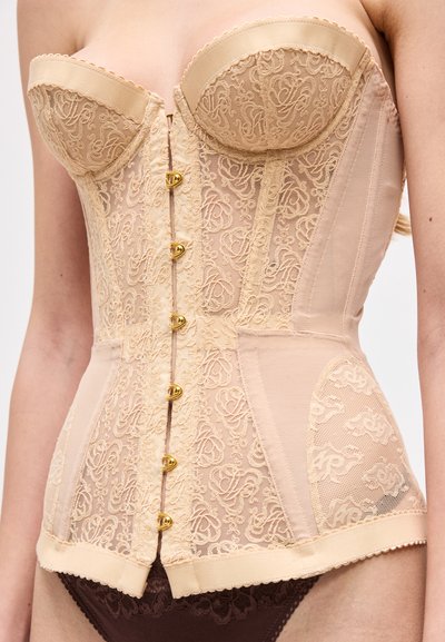 Μπεζ κορσές από δαντέλα με δομημένα cups και χρυσά κλείστρα με αγκράφες, φορεμένος πάνω από καφέ δαντελένιο εσώρουχο στο κορμί ενός μοντέλου.