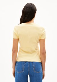 Žluté žebrované tričko s krátkým rukávem a kulatým výstřihem, zobrazené zezadu. Kombinováno s modrými džíny s vysokým pasem.