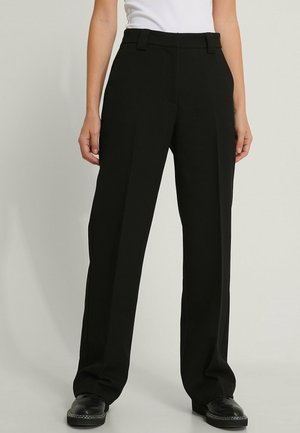 Mujer con pantalones negros de corte recto con trabillas para cinturón y zapatos negros, combinados con una camiseta blanca metida por dentro.