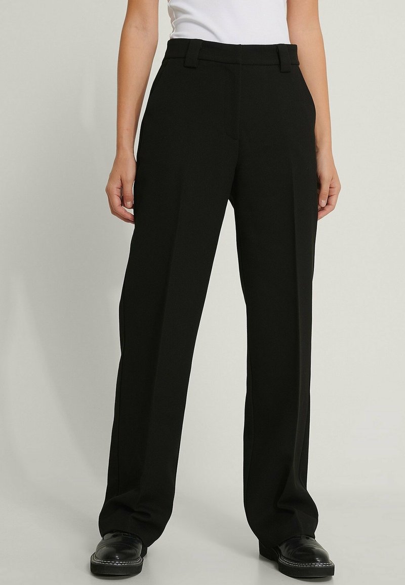 Femme portant un pantalon noir droit avec des passants pour ceinture et des chaussures noires, associé à un haut blanc rentré.