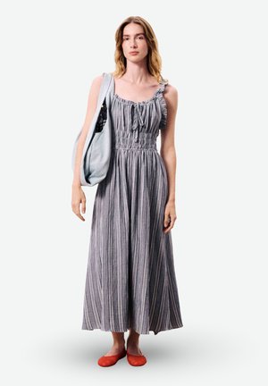 Femme portant une robe maxi grise à rayures sans manches, des ballerines rouges et un sac à épaule gris clair sur son épaule gauche.