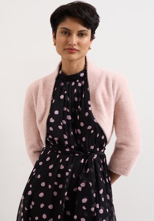 Femme aux cheveux courts et foncés portant des boucles d’oreilles créoles dorées, un châle duveteux rose clair et une robe noire à pois roses avec une ceinture à la taille.