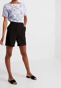 Svetle modrá květinová blůza s krátkými rukávy, doplněná černými přiléhavými šortkami. Oblečení je doplněno černými sandály na slip-on.