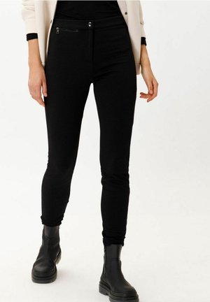 Kvinde iført teksturerede sorte slimfit bukser, sorte støvler og en beige blazer med sorte manchetter, stående mod en hvid baggrund.