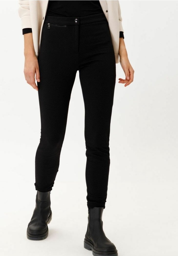 Femme portant un pantalon slim noir texturé, des bottes noires et un blazer beige avec des poignets noirs, debout sur un fond blanc.