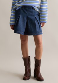 Gonna di denim con pieghe, lavaggio blu e cuciture a contrasto, abbinata a stivali alla caviglia in pelle marrone e un maglione a righe.