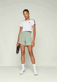 Sieviete stāv sporta apģērbā, turēt melnu ūdens pudeli, ģērbusies baltā īsajā krekliņā, gaiši zaļās gumijas vidukļa šortos, baltās zeķēs un kedās.