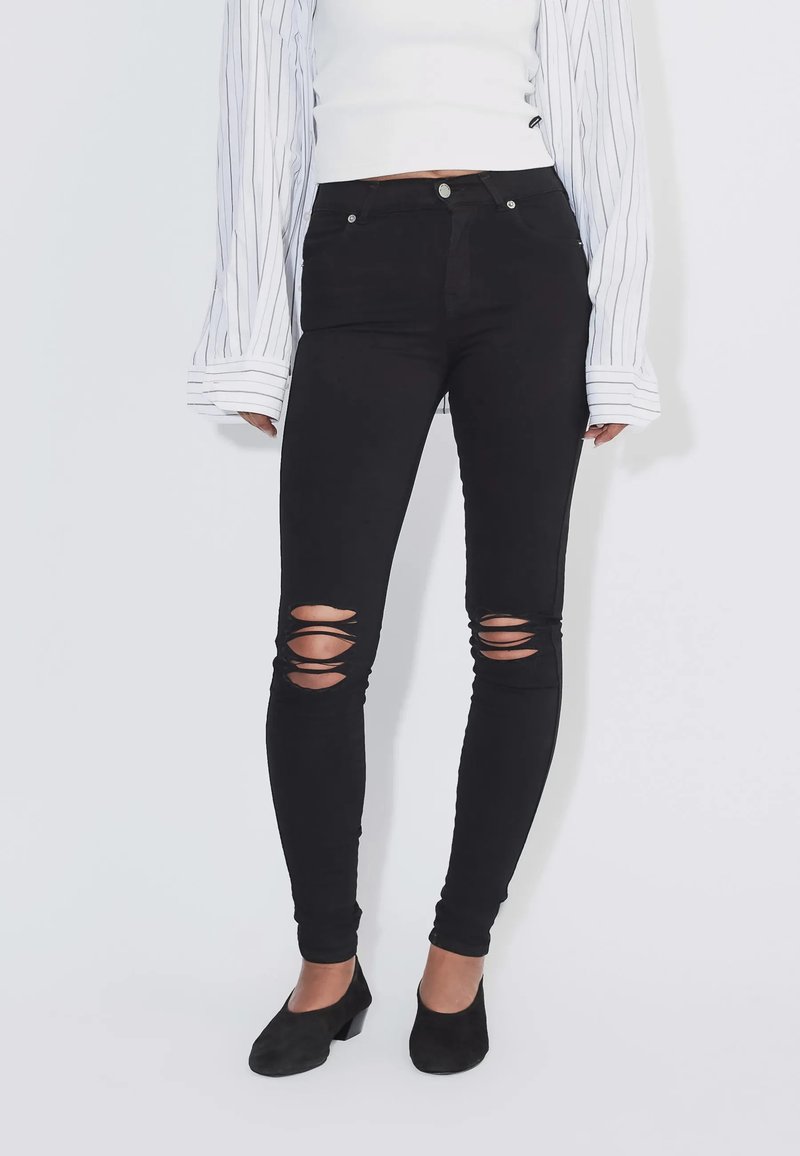 Persona che indossa jeans skinny neri strappati, scarpe nere con tacco, un top corto bianco e una camicia bianca a maniche lunghe con righe nere.