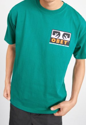 Grünes Baumwoll-T-Shirt mit einem grafischen Logo, das schwarze und weiße Hände zeigt, sowie orangem 'OBEY'-Text auf der Brust. Kurze Ärmel, lockere Passform.