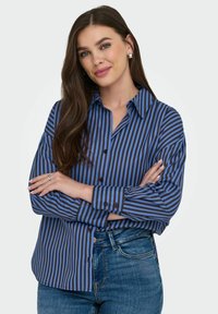 Blouse rayée bleu et noir avec un devant à boutons, un col pointu et des manches longues. Le tissu semble lisse. Associée à un jean en denim.