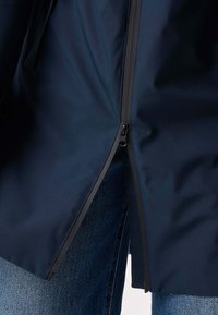 Mørkeblå jakke med sort lynlås delvist åbnet, der afslører blå denimjeans under, vist fra lårets niveau.