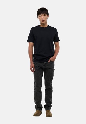 Jeune homme debout avec une main dans la poche, portant un t-shirt noir, un jean foncé et des bottes en daim marron sur fond blanc.