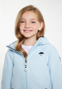 Ljusblå zip-hoodie i texturerat material, med huva, framfickor och en liten svart paraplylogotyp på bröstet.
