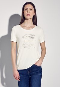 Krémové tričko s krátkým rukávem, které má metalický nápis na přední straně, kulatý výstřih a měkkou tkaninu. V kombinaci s modrými džíny.