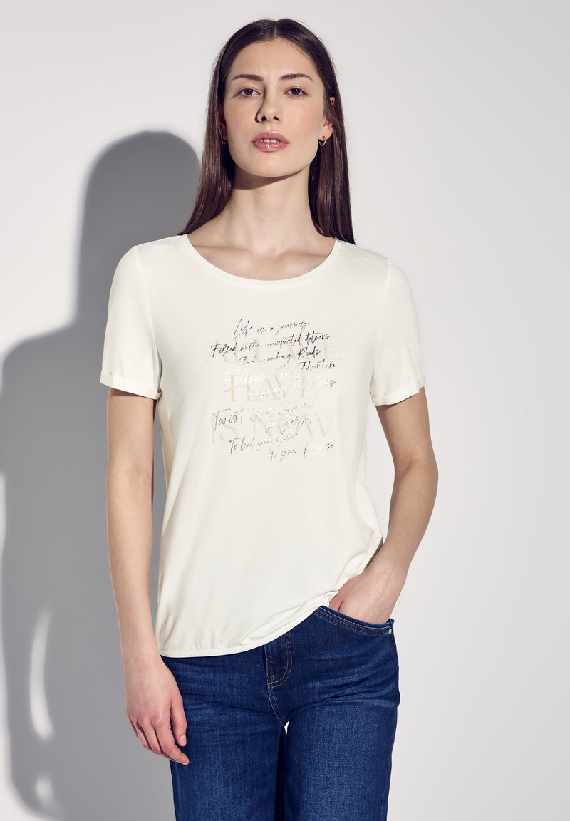 Krémové tričko s krátkým rukávem, které má metalický nápis na přední straně, kulatý výstřih a měkkou tkaninu. V kombinaci s modrými džíny.