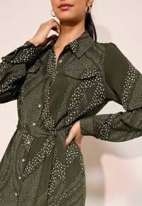 Rochie cambrată de culoare verde măslinie dintr-un material moale, cu un model cu puncte albe, cu deschidere la nasturi, două buzunare pe piept și o curea asortată din același material.