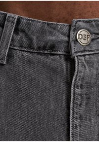 Pantalones de mezclilla gris con cintura plana, costuras visibles y un botón metálico que lleva el texto "DEF." Tejido texturizado y color apagado.