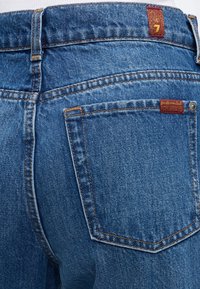 Πίσω τσέπη από μπλε τζιν με πορτοκαλί ραφές και μικρές κόκκινες ετικέτες, η μία με τον αριθμό επτά και η άλλη με κείμενο στην άκρη της τσέπης.