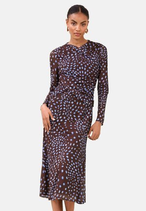 Nő hosszú ujjú, barna midi ruhában, világoskék absztrakt pöttyös mintával, kezét a csípője közelében tartva, egyszínű háttér előtt pózol.