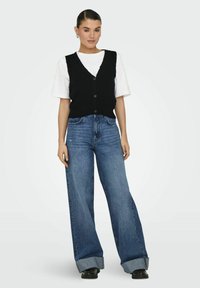 Schwarze gestrickte Weste mit Knöpfen, getragen über einem weißen T-Shirt. Kombiniert mit hellblauen weit geschnittenen Jeans und schwarzen Schuhen. Glatte Stofftextur.