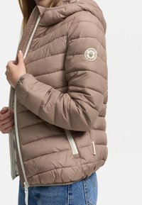 Brun pufferjacka med huva, med horisontellt quiltat mönster, vit dragkedja och en logopatch på vänster ärm.